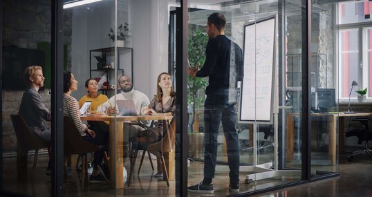 Project Manager Makes a Presentation for a Young Diverse Creative Team in Meeting Room in an Agency. Colleagues Sit Behind Conference Table and Discuss Business Development, User Interface and Design. 