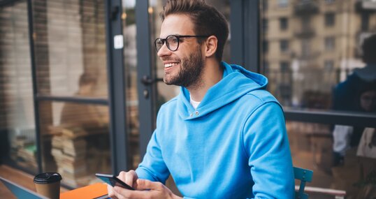 Portrait of cheerful millennial blogger with modern cellphone and laptop technology enjoying freelance lifestyle, happy hipster guy in optical eyewear using mobile phone and netbook in street cafe