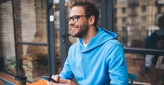 Portrait of cheerful millennial blogger with modern cellphone and laptop technology enjoying freelance lifestyle, happy hipster guy in optical eyewear using mobile phone and netbook in street cafe
