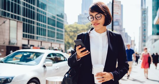 Concentrated female entrepreneur using application for calling taxi getting to work with morning coffee to go, prosperous businesswoman checking notification on smartphone strolling with takeaway.