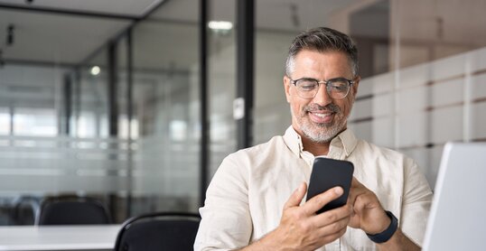 Middle aged Hispanic business manager ceo using cell phone mobile app, laptop. Smiling Latin or Indian mature man businessman holding smartphone sit in office working online on gadget with copy space.