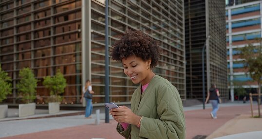 Sideways shot of beautiful woman with Afro hair holds smartphone reads received sms message has cheerful expression sits in downtown against cityscrapers dials number of taxi service. Technology