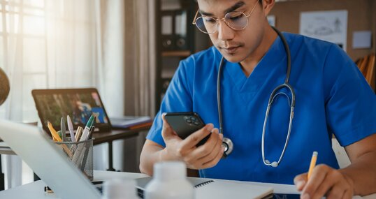 Mid adult male doctor reviews patient records on smartphone and desktop PC.