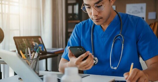 Mid adult male doctor reviews patient records on smartphone and desktop PC.