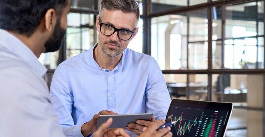 Diverse managers traders analysts discussing financial growth market at desk with laptop with graphs on screen using tablet device. Investors brokers analysing indexes online cryptocurrency stock