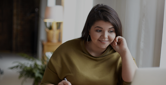 Modern electronic gadgets, job and occupation concept. Cute elegant young brunette female with excess weight using laptop computer for remote work, looking at screen, holding pen, writing down