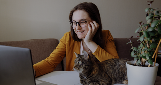 Remote work. The girl is sitting at the computer and looks at screen. A tabby cat sits next to me and also looks at screen. On a white table is a potted flower. A person sincerely smiles at someone. 